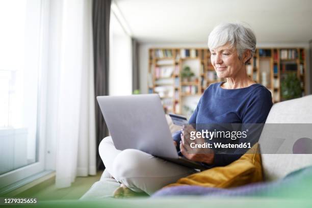 senior woman using credit card while doing online shopping on laptop at home - silver surfer - fotografias e filmes do acervo