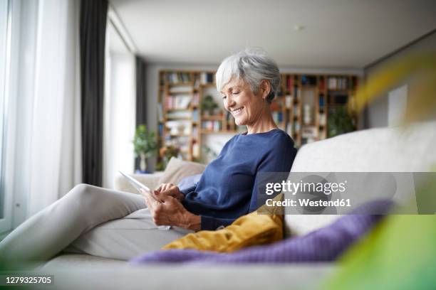 smiling woman looking in digital tablet while sitting at home on sofa - old livingroom stock-fotos und bilder