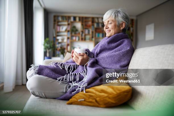 thoughtful senior woman with blanket holding coffee cup while sitting on sofa at home - blanket photos et images de collection