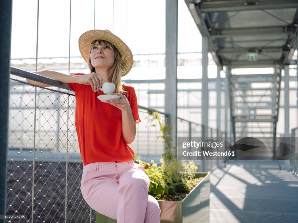 Thoughtful businesswoman wearing hat holding coffee cup while sitting by railing in greenhouse