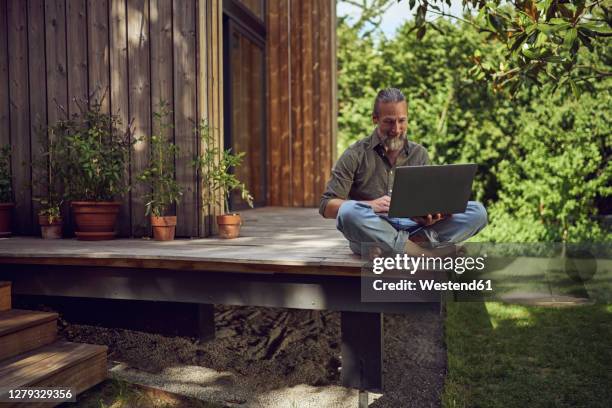 mature man using laptop while sitting against tiny house - grounds worker stock pictures, royalty-free photos & images