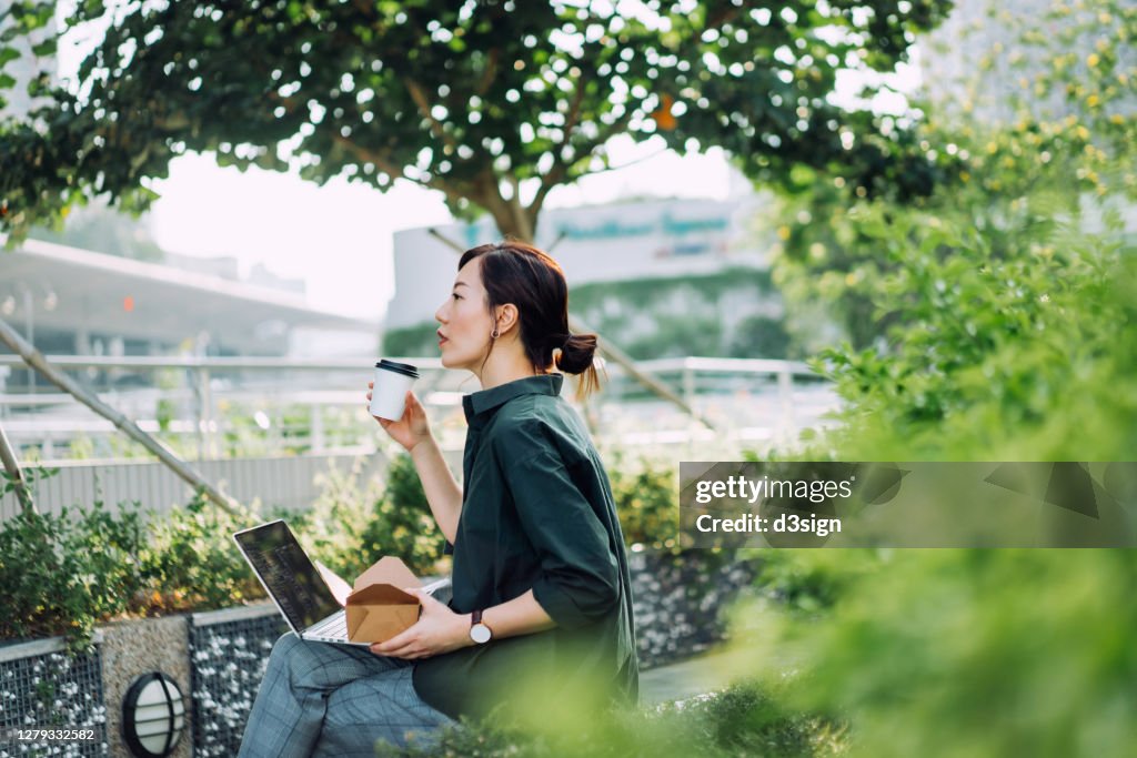 Professional young Asian businesswoman sitting on the bench in an urban park, working on laptop while having a healthy salad lunch box with a cup of coffee during lunch break