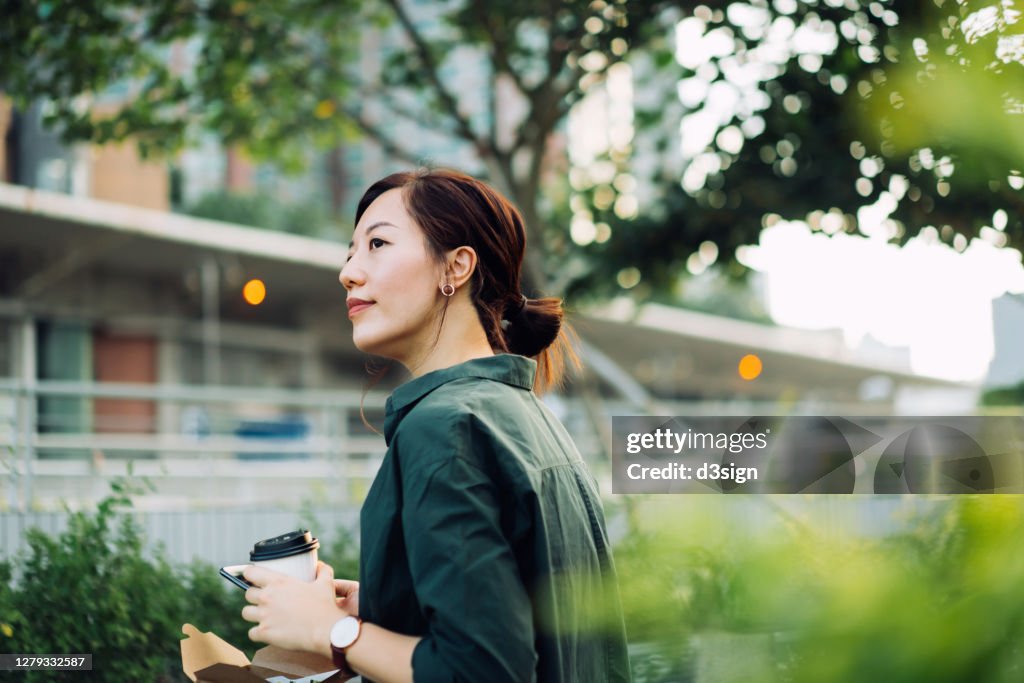 Smiling young Asian businesswoman using smartphone while having a healthy salad lunch box with a cup of coffee outdoors in an urban park during lunch break