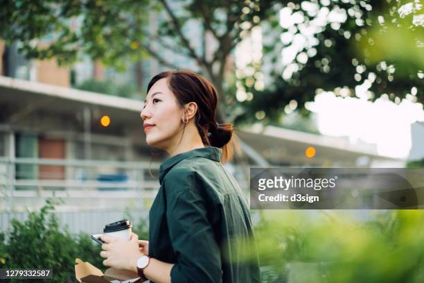 smiling young asian businesswoman using smartphone while having a healthy salad lunch box with a cup of coffee outdoors in an urban park during lunch break - city life stock pictures, royalty-free photos & images