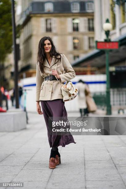 Model wears a beige double breasted jacket, a large belt, a bag, a purple lustrous silky flowing dress, black tights, brown leather shoes, outside...