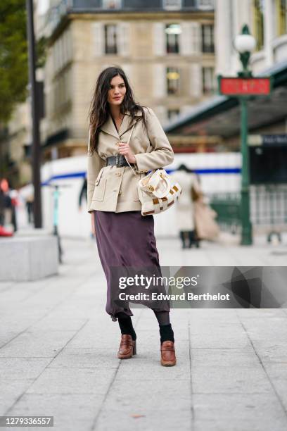 Model wears a beige double breasted jacket, a large belt, a bag, a purple lustrous silky flowing dress, black tights, brown leather shoes, outside...