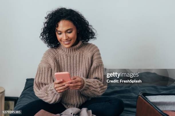 happy young woman packing her suitcase for winter holiday and booking a flight online on her mobile phone - phone mid air stock pictures, royalty-free photos & images