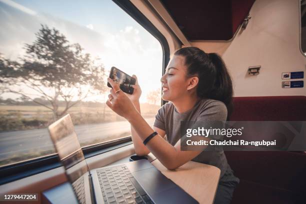 asian teen is a backpacker traveling by train, asia tourist woman take a photo by smart phone out the window with a notebook laptop on the table - vacation train stock pictures, royalty-free photos & images