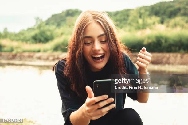 giovane donna sorridente seduta accanto a un fiume digitando messaggi sul suo smartphone - eccitazione foto e immagini stock