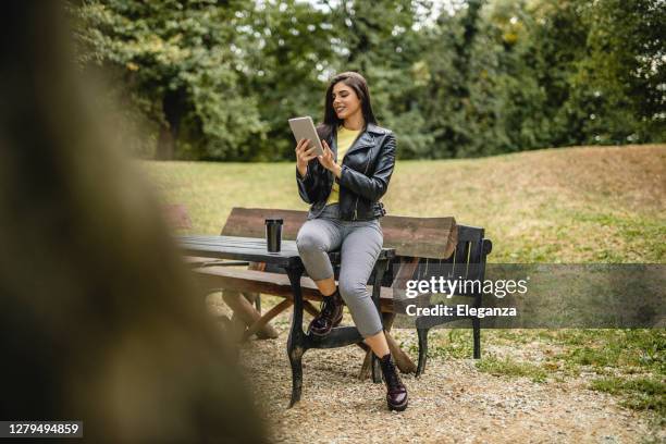 young beautiful woman drinking coffee and reading e-book on digital tablet outdoors - nature magazine stock pictures, royalty-free photos & images