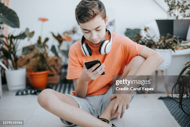 teenage boy with headphones using mobile phone at table in room. 11 years old boy sitting behind a laptop and listening to music with headphones or playing video game. - 12 years stock-fotos und bilder