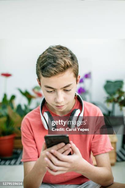 teenage boy with headphones using mobile phone at table in room. 11 years old boy sitting behind a laptop and listening to music with headphones or playing video game. - 12 years stock-fotos und bilder