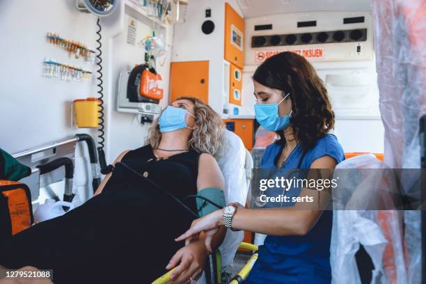 female paramedic with face mask helping a patient and measures patient’s blood pressure in ambulance during pandemic - heart attack stock pictures, royalty-free photos & images