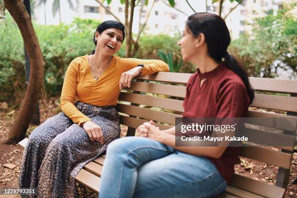 happy looking sisters discussing while sitting on a bench in public park - chatting park stockfoto's en -beelden