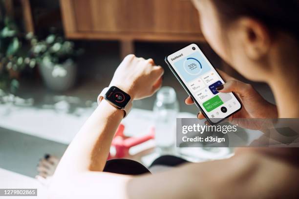 over the shoulder view of young asian sports woman checking pulse on smartwatch and syncing her smartwatch with fitness app on smartphone to monitor her training progress after working out / exercising / practicing yoga at home in the fresh bright morning - smart watch stock pictures, royalty-free photos & images