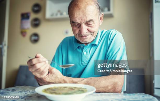 elderly man holds his hand trying to control parkinson disease while eating a soup. - spoon in hand stock-fotos und bilder