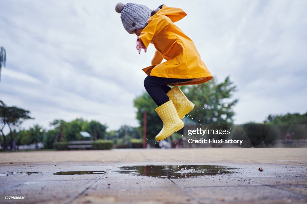 Mid-air shot of a child jumping in a puddle of water wearing yellow rubber boots and a raincoat in autumn