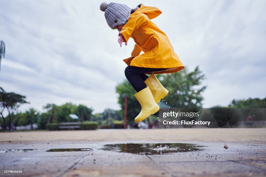 秋天, 一個孩子穿著黃色橡膠靴和雨衣在水坑裡跳的半空中鏡頭。