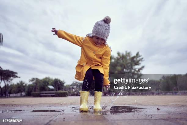 chica sonriente linda salpicando agua en un charco en botas de goma amarillas - abrigo amarillo fotografías e imágenes de stock