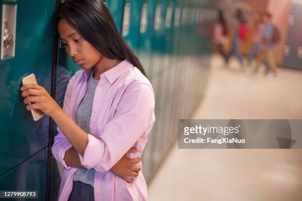 lonely teenage asian female student leaning on locker with smart phone in school - cyberbullying stock pictures, royalty-free photos & images