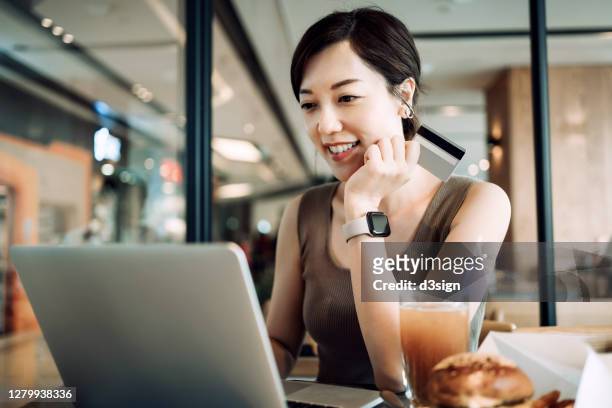 beautiful smiling young asian woman having meal in a cafe, shopping online with laptop and making mobile payment with credit card on hand - asian credit card imagens e fotografias de stock