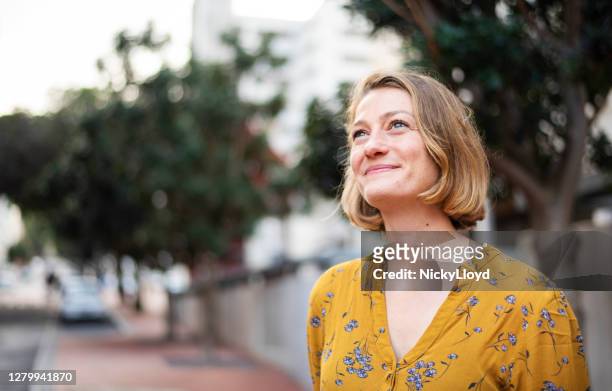 smiling young woman looking up while standing on a city sidewalk - looking up stock pictures, royalty-free photos & images