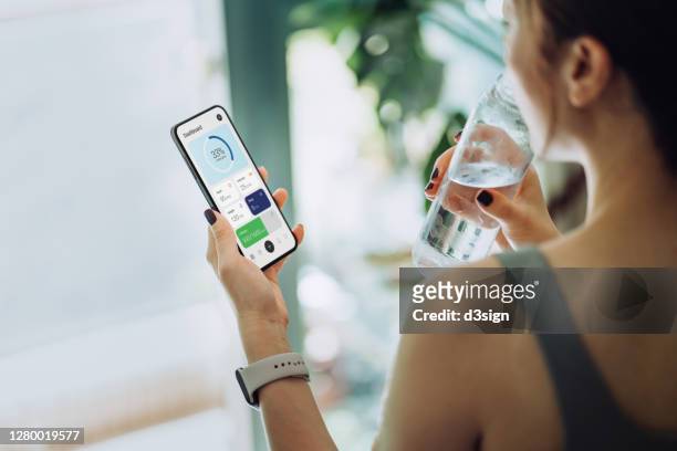 over the shoulder view of young asian sports woman refreshing with water and using fitness app on smartphone to monitor her training progress after fitness work out / exercising / practicing yoga at home in the fresh bright morning - application performance stock-fotos und bilder