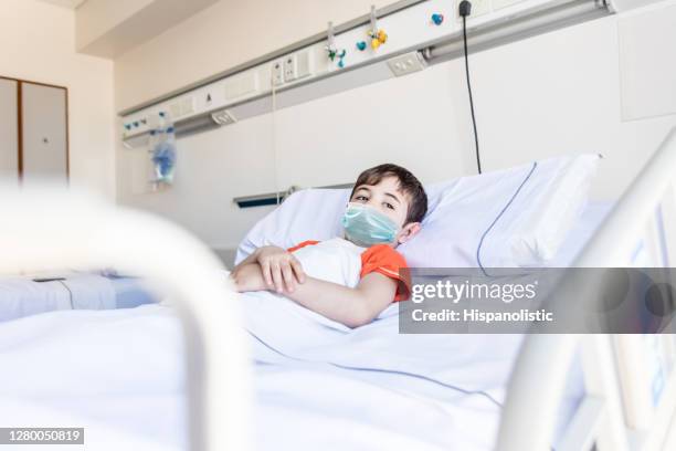 pensive little boy lying down on hospital bed looking away with sadness wearing a protective facemask - child lying down stock pictures, royalty-free photos & images