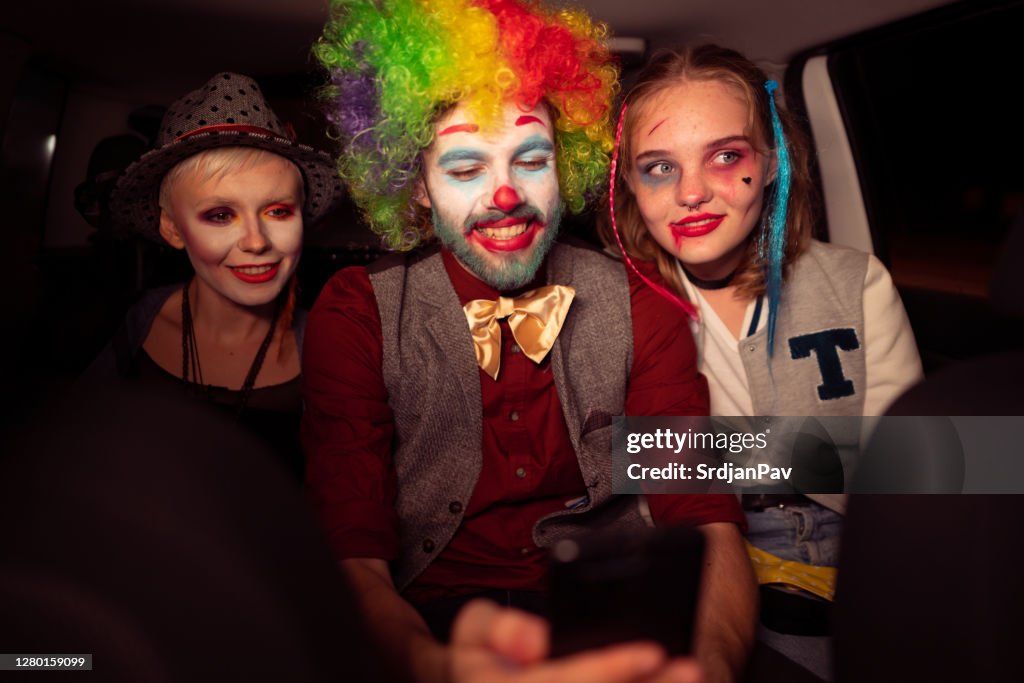 Freunde in Kostümen, fahren im Auto in der Halloween-Nacht, während ein junger Mann sein Handy benutzt