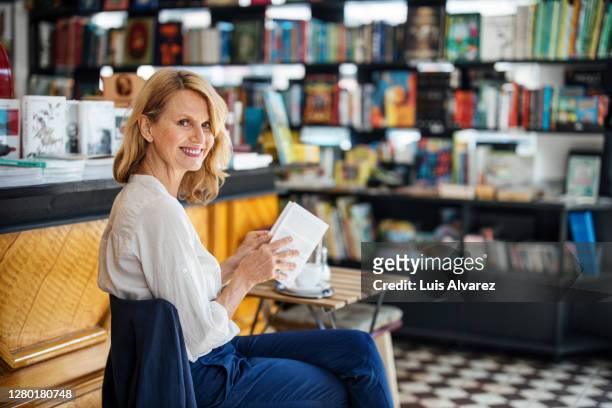 smiling mature woman in cafe with a book - bookstore stock-fotos und bilder