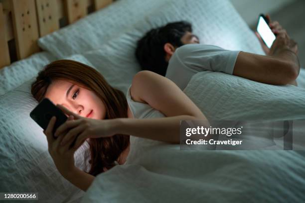 shot of a young couple using their cellphones in bed at morning back to back - couple ignore stock-fotos und bilder