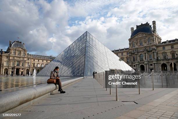 Woman wearing a protective face mask seats in front of the empty courtyard of the Louvre museum and the pyramid of Louvre without tourists on October...