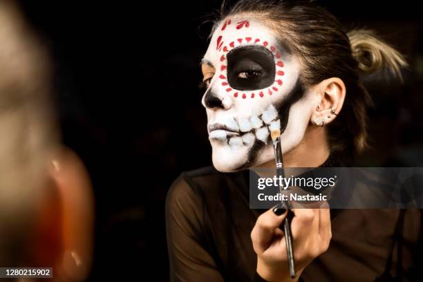 young woman painting face for halloween night party looking at mirror holding make up pencil - face painting stock pictures, royalty-free photos & images