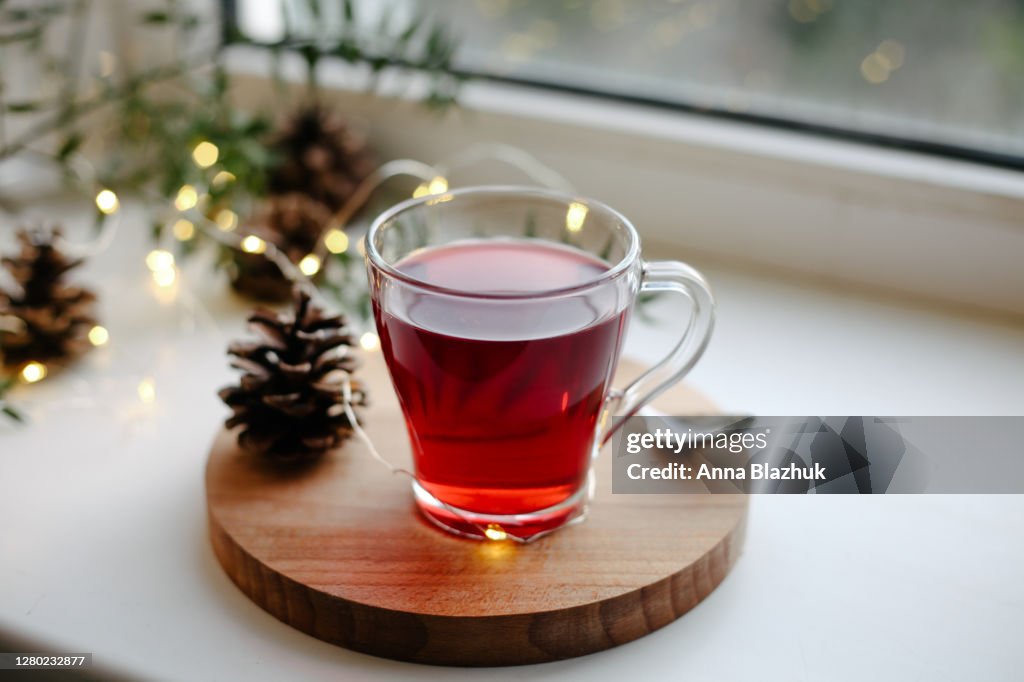 Hot homemade winter drink in glass cup on window sill, pine cones and Christmas decorations