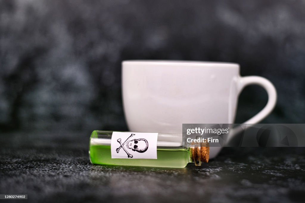Vial with green liquid and poison skull label in front of blurry tea cup on dark background