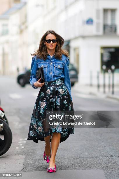 Therese Hellström wears sunglasses, a blue denim jacket from Custommade, a studded black leather clutch from Valentino, a black pleated floral print...