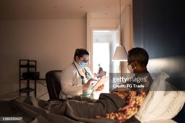 Médico hablando con la paciente de la tercera edad en la visita médica en su casa - usando máscara facial