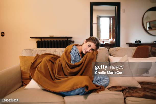 young woman wearing pajamas and a blanket sitting on sofa - autumn living room stock pictures, royalty-free photos & images