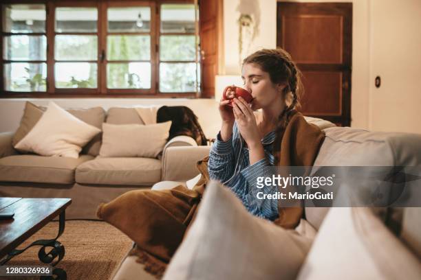 young woman drinking from a cup while sitting on sofa - roupa quente imagens e fotografias de stock