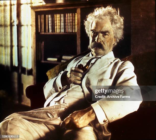 Colorized portrait of American author and humorist Mark Twain, born Samuel Langhorne Clemens holding a pipe and glasses, with a newspaper in his lap,...