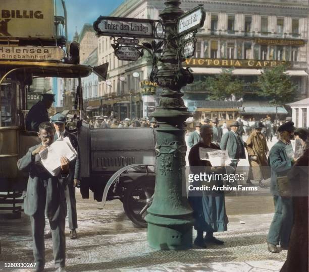 Colorized photo of a street scene at the intersection of Unter den Linden and Friedrichstrasse, Berlin, Germany, 1907. A newsvendor sells papers to...