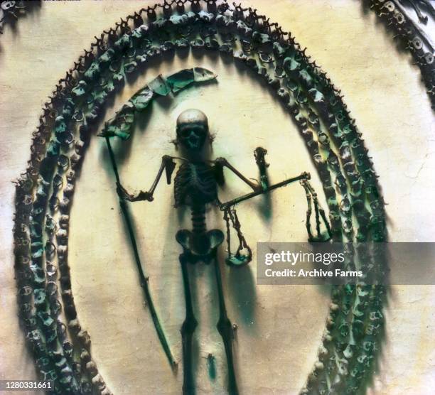 Colorized photo of a skeleton, holding a scythe and a scale, enclosed in an oval of bones inside the Crypt of the Three Skeletons, one of the rooms...