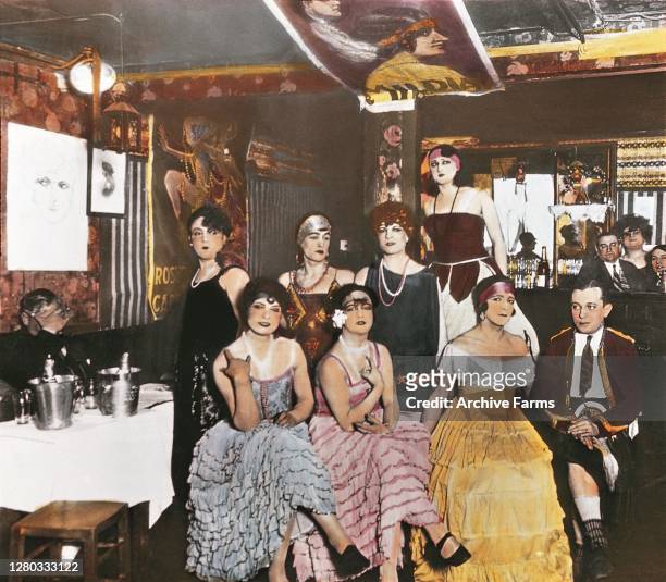 Colorized photo of patrons, a number of whom wear elaborate evening gowns, in a bar in Montmartre, Paris, France, 1927. The original caption contains...