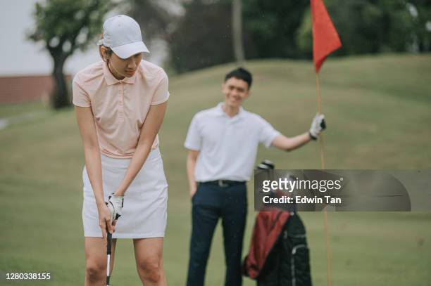 asian chinese female golfer putting ball with male partner holding flag on golf course - golf lessons stock pictures, royalty-free photos & images