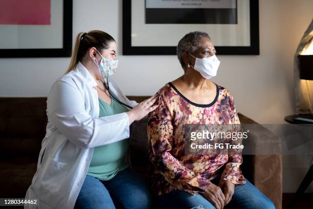 Doctor using stethoscope listening to senior patient breathing at her house - using face mask