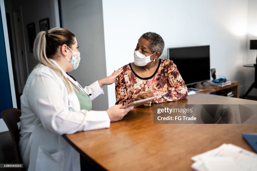 Doctor talking to senior woman patient on medical visit at her home - using face mask