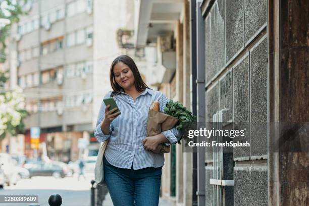 mujer de tamaño más sonriente lleva una bolsa de papel llena de comida y usando su teléfono móvil en la calle - woman carrying tote bag fotografías e imágenes de stock