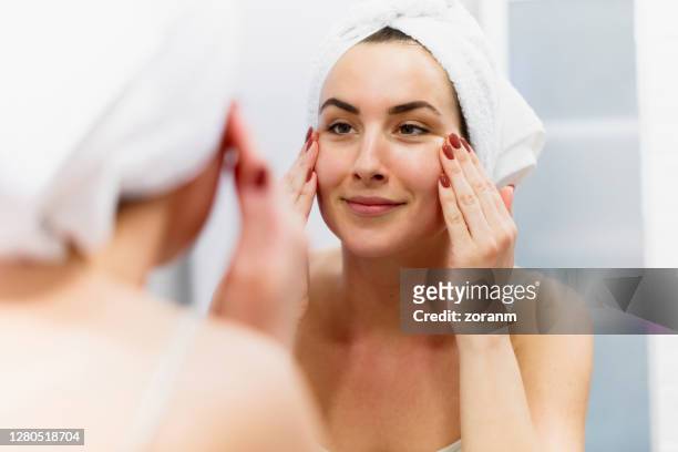 smiling woman wearing towel on wet hair tightening her skin in front of the mirror - face lift stock pictures, royalty-free photos & images