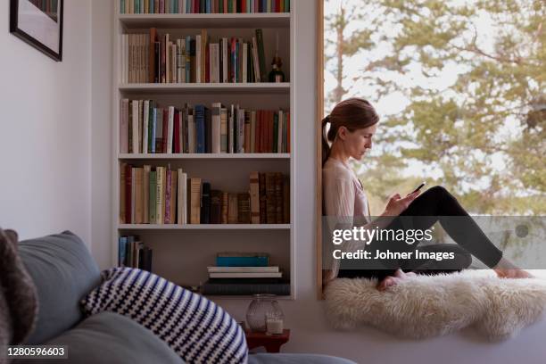 woman at home using cell phone - bookshelf stockfoto's en -beelden