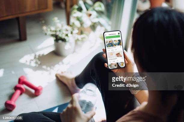 over the shoulder view of young asian sports woman using meal delivery service and ordering healthy food online with smartphone after exercising at home - sportswear shopping stock-fotos und bilder